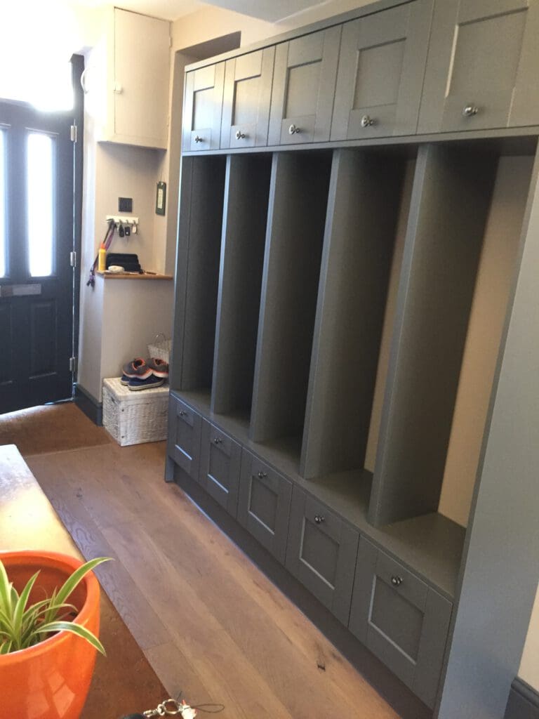 A hallway with light wooden flooring features a row of dark green closed cubby storage units on the right. The wall has hooks holding keys and sunglasses. A small table with an orange plant pot is in the foreground. A black front door is partially visible.