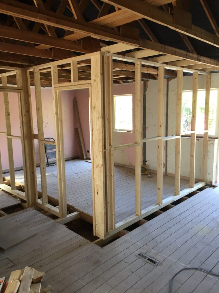 A room under construction with wooden framing and exposed studs. The floor is made of wooden planks, and the walls lack drywall. Sunlight streams in through unfinished window frames. Tools and construction materials are scattered around.