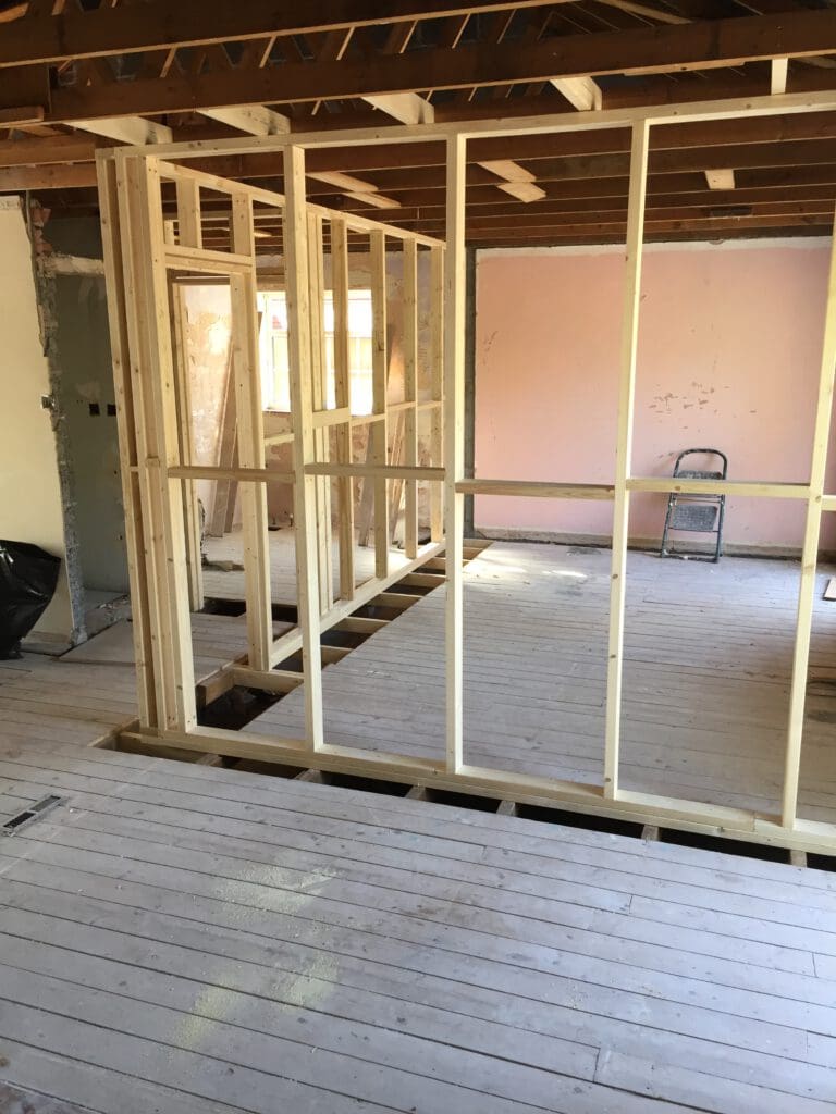 Partially constructed interior wall with exposed wooden studs in a room undergoing renovation. The floor is wooden, walls are partially unfinished, and the ceiling shows exposed beams. A ladder is positioned against a pink wall, and some tools and debris are scattered around.