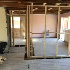 An unfinished room with exposed wooden framing and a partially constructed wall. The floor is bare with scattered construction materials. There's a ladder against one wall, and sunlight streams in through the windows in the background.