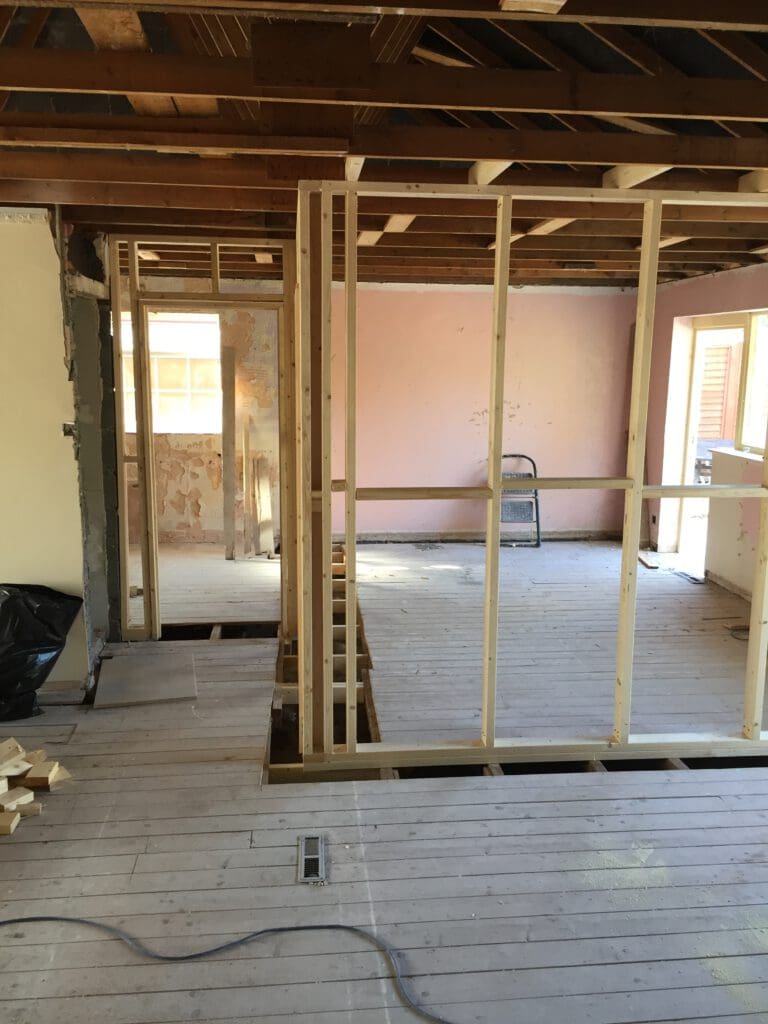 An unfinished room with exposed wooden framing and a partially constructed wall. The floor is bare with scattered construction materials. There's a ladder against one wall, and sunlight streams in through the windows in the background.