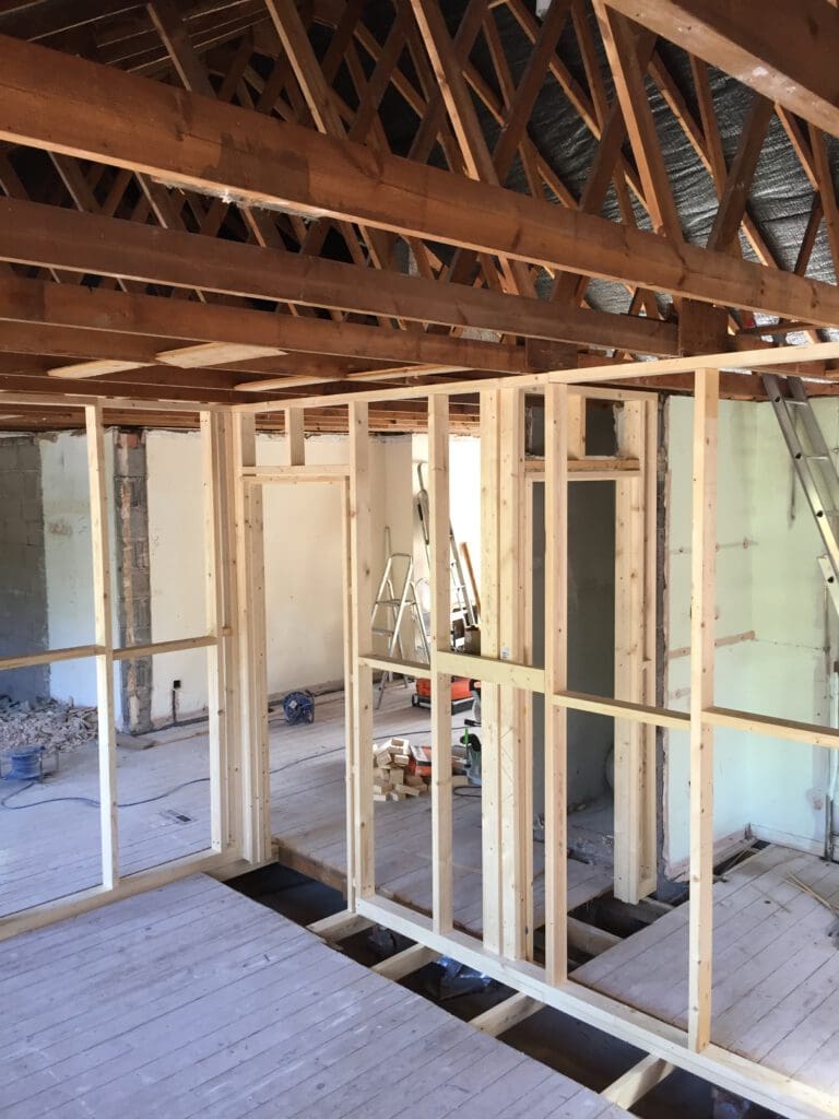 A partially constructed interior showing wooden framing for walls and ceiling joists. There are tools, a ladder, and building materials scattered around the space. The floor is made of wooden planks, and the ceiling has exposed beams.