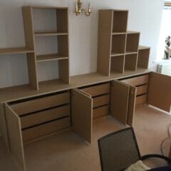 A home office in progress with unfinished wooden shelving units, including both open shelves and cabinets with open doors, lining a wall. There is a small dining table with a chair in the foreground and a window on the right side.