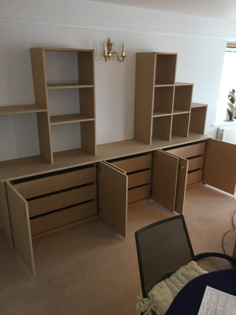 A home office in progress with unfinished wooden shelving units, including both open shelves and cabinets with open doors, lining a wall. There is a small dining table with a chair in the foreground and a window on the right side.