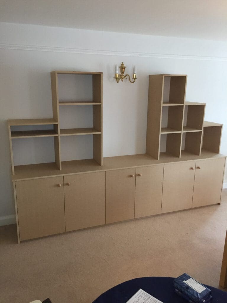 A wooden cabinet setup against a white wall featuring multiple shelves and closed compartments at the bottom. The unit is topped with open shelves of varying heights. Above the cabinet is a brass wall-mounted light fixture with white shades.