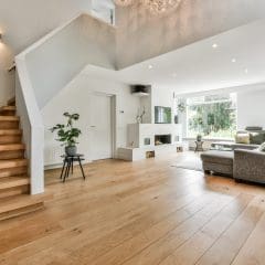 Modern living room with wooden floors, a grey sectional sofa, and a minimalistic design. The room features a wooden staircase with white railings, indoor plants, and large windows allowing ample natural light to brighten the space.