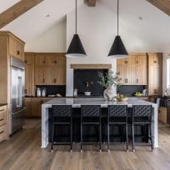 A modern kitchen with wooden cabinets and a vaulted ceiling features a black countertop island with wicker barstools. Two large pendant lights hang above. A stainless steel fridge, window, and black accents complete the design.