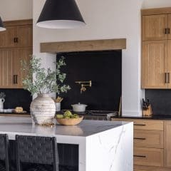 Modern kitchen with light wooden cabinets, a large white marble island, and black leather barstools. A vase with greenery and a bowl of green apples sit on the island. Two black pendant lights hang above.